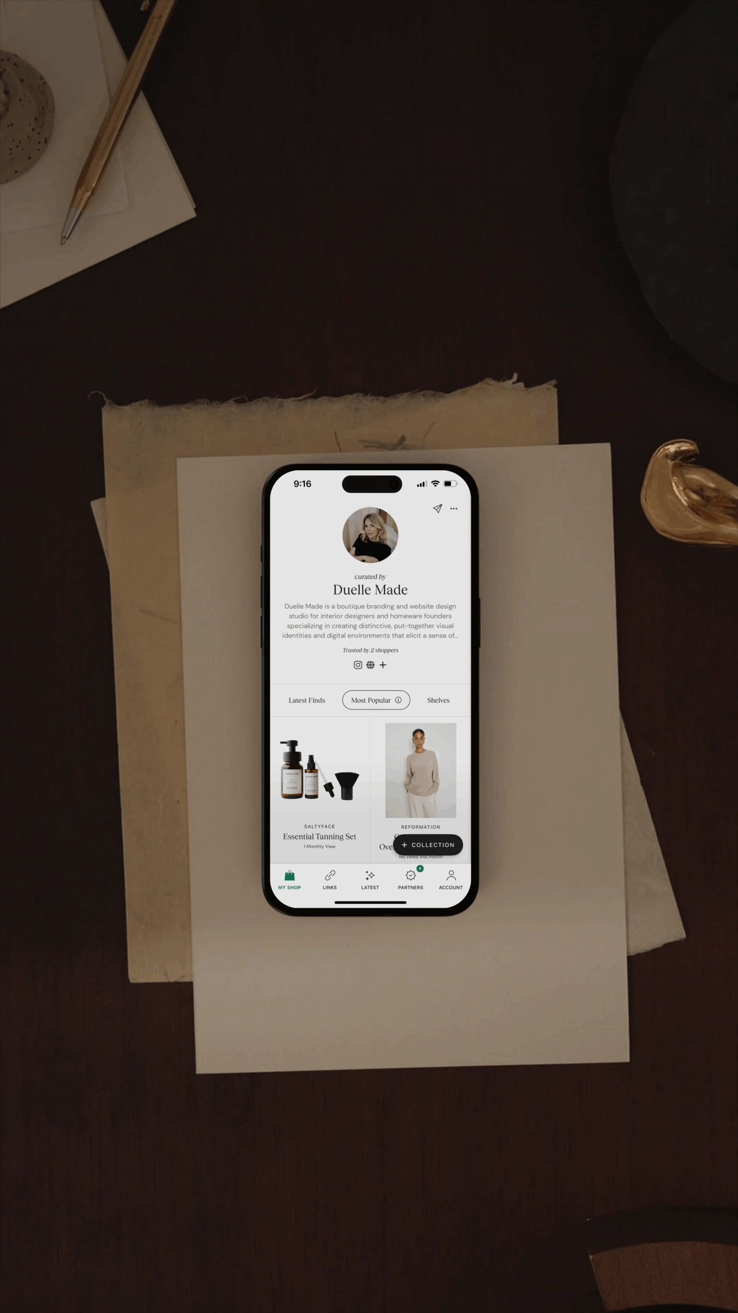 A smartphone displaying Duelle Made’s ShopMy profile stands upright on a wooden surface beside a lamp in a softly lit, neutral-toned room. The screen shows the Duelle Made profile description and curated product collections, with a warm and minimal aesthetic.