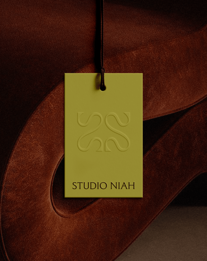 Close-up of a product hangtag for Studio Niah. The olive green tag features a blind-embossed logo and rests against a rust-colored velvet fabric, highlighting the brand's focus on rich textures.