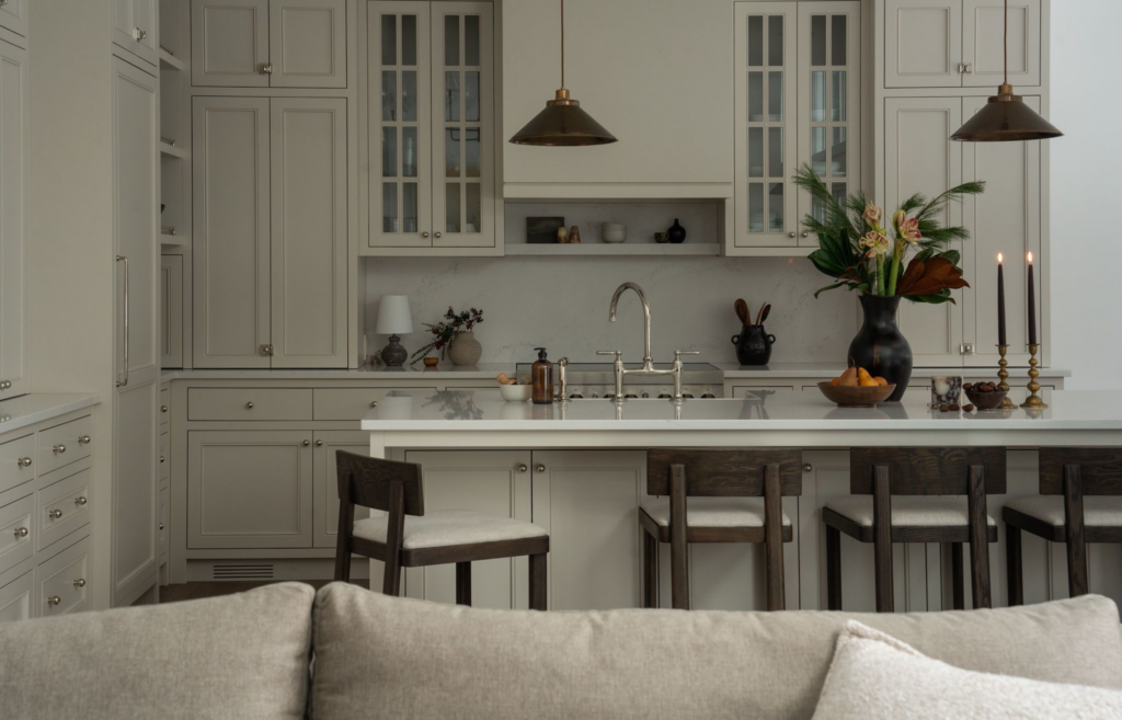 A wide shot of a spacious, elegant kitchen with warm-grey traditional cabinetry and a large central island topped with white marble. Four dark wood counter stools with cream-cushioned seats are lined up at the island. The island is decorated with a tall black ceramic vase holding winter evergreens and amaryllis flowers, a wooden bowl of pears, and two tall brass candlesticks with lit dark taper candles. Above the island hang two cone-shaped dark bronze pendant lights, and a classic gooseneck faucet sits by a small white prep sink. The top of a neutral linen sofa is visible in the immediate foreground, framing the view into the kitchen.