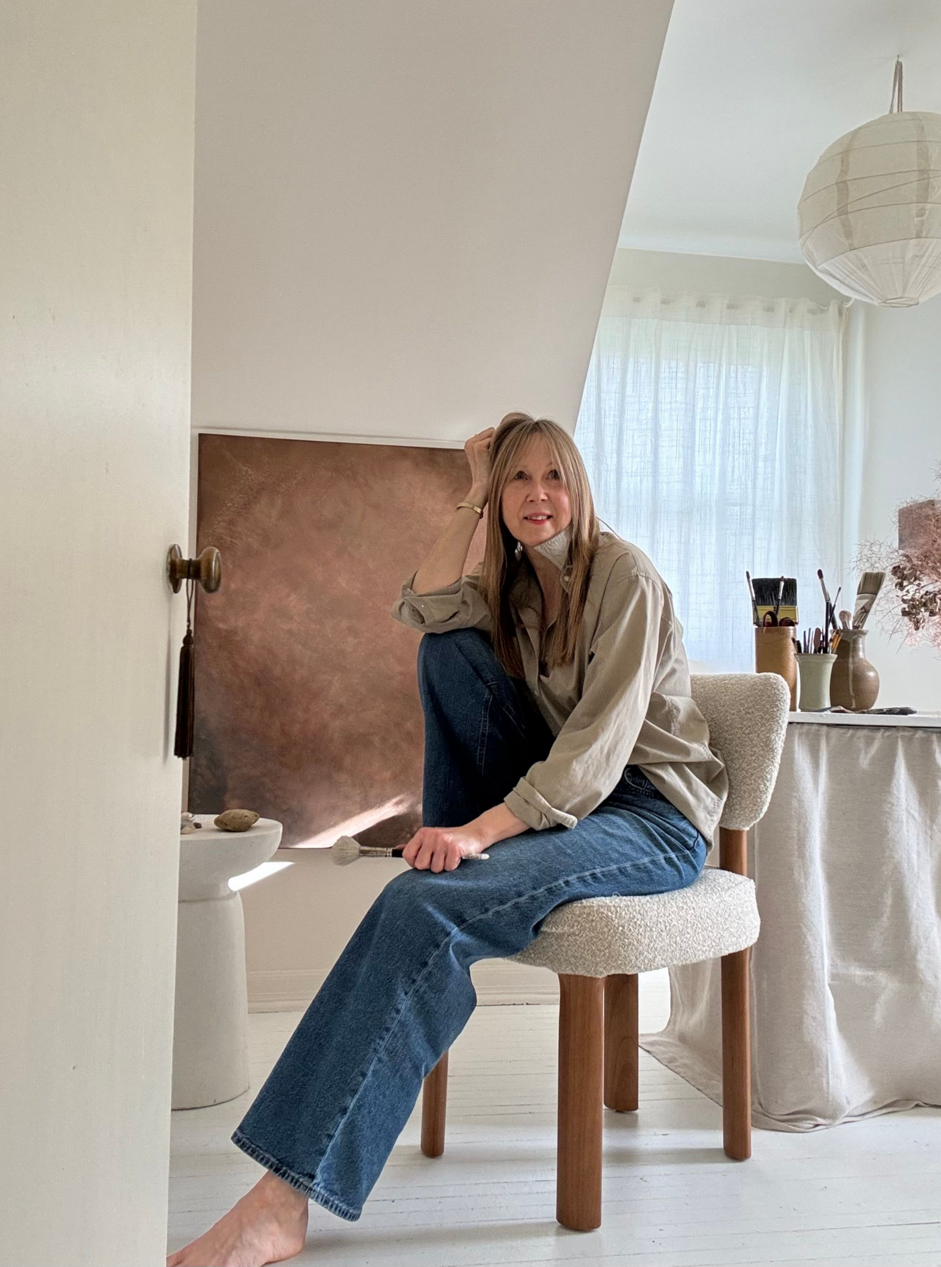 A full-frame portrait of a woman with long brown hair sitting barefoot on a textured cream bouclé chair in a sunlit artist's studio. She is dressed casually in a beige button-down shirt and blue jeans, holding a paintbrush and looking off-camera with a gentle smile. Behind her, a large abstract canvas painted in rich, metallic bronze and brown tones rests against a white wall under a sloped ceiling. To the right, a draped worktable holds jars overflowing with artist brushes beneath a large, round paper lantern pendant light.