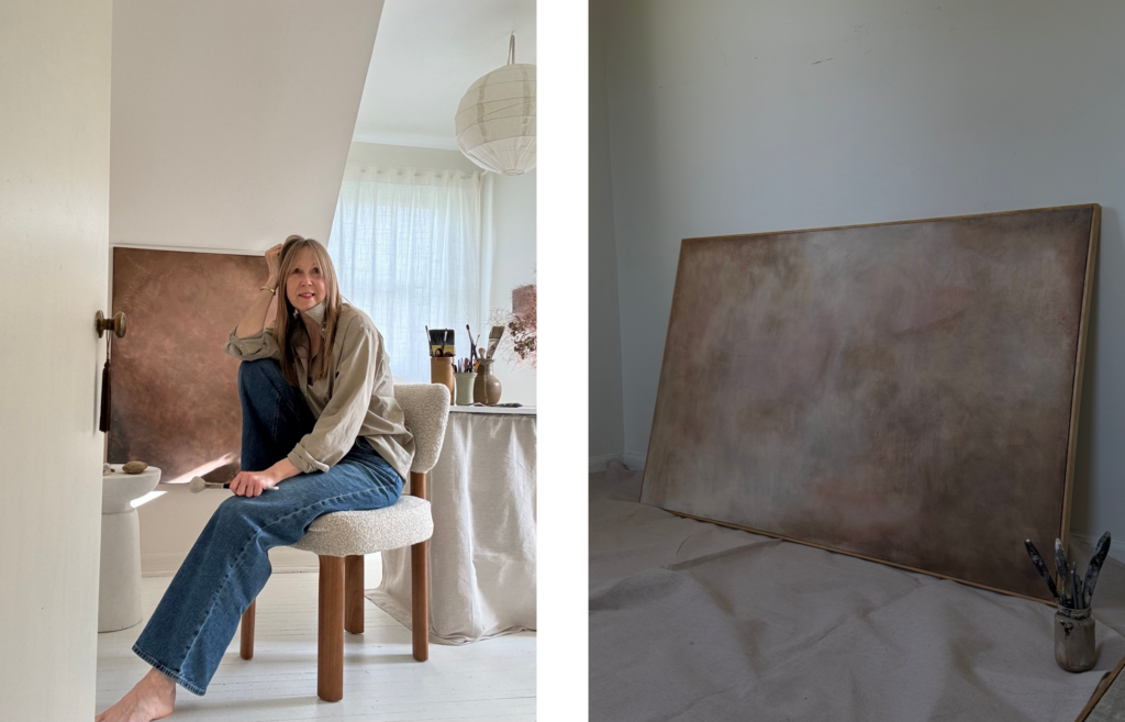 Left side: A woman with long brown hair sits barefoot on a textured cream-colored chair in a bright artist's studio. She wears a beige button-down shirt and blue jeans, holding a paintbrush and looking thoughtfully to the side. In the background, a large earthy, bronze-toned abstract painting rests against the wall under a sloped ceiling, next to a table filled with paintbrushes.
Right side: A large, unframed abstract painting with soft, textured shades of brown, beige, and muted pink rests on a canvas drop cloth against a white wall. A small glass jar holding a few paintbrushes sits in the lower right corner.