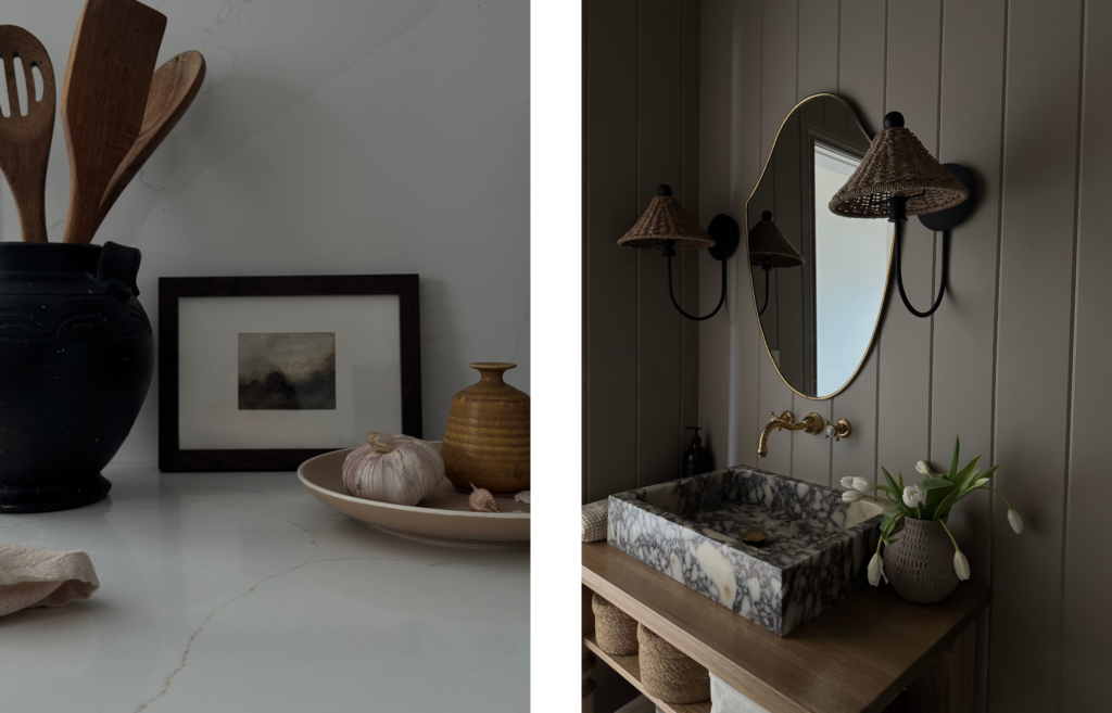 Left side: A moody kitchen still life on a white marble countertop. On the left, a large matte black ceramic jar holds wooden cooking utensils. Next to it sits a small, dark-framed landscape painting, a beige ceramic dish with a head of garlic, and a small mustard-yellow textured vase.
Right side: A minimalist bathroom vanity featuring a rectangular sink carved from heavily veined grey and white marble. A curved gold faucet is mounted to the beige tongue-and-groove paneled wall beneath an oval brass-rimmed mirror. Flanking the mirror are two black wall sconces with woven rattan shades, and a small vase of white tulips sits on the wooden vanity.