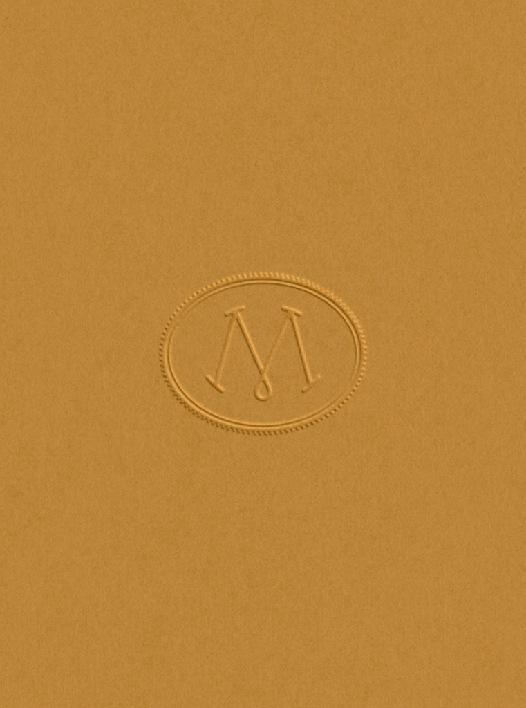 A close-up of a textured, mustard-yellow surface featuring a subtle, blind-embossed logo in the center. The logo consists of an elegant, intertwining letter "M" enclosed within a classic oval border with a rope-like texture.