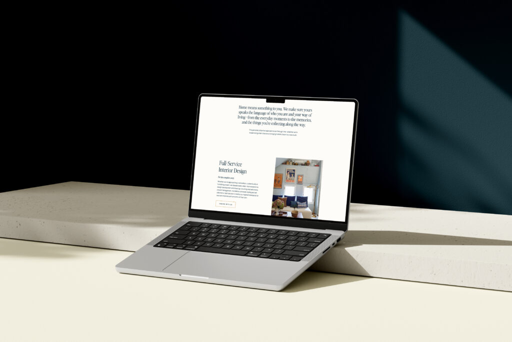 An open silver laptop sitting on a minimalist cream-colored desk surface against a stark black background. The laptop screen displays a clean, white-background website for interior design, featuring a large text block at the top and a photo of a cozy, well-decorated room below the header "Full Service Interior Design".
